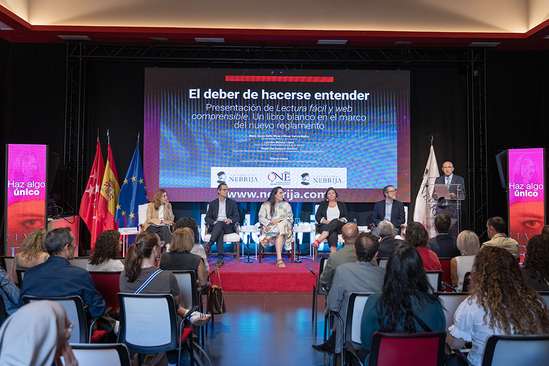 La mesa redonda en el escenario: cinco participantes sentados, un orador en el atril y el público en primer plano. En la pantalla, el título «El deber de hacerse entender».