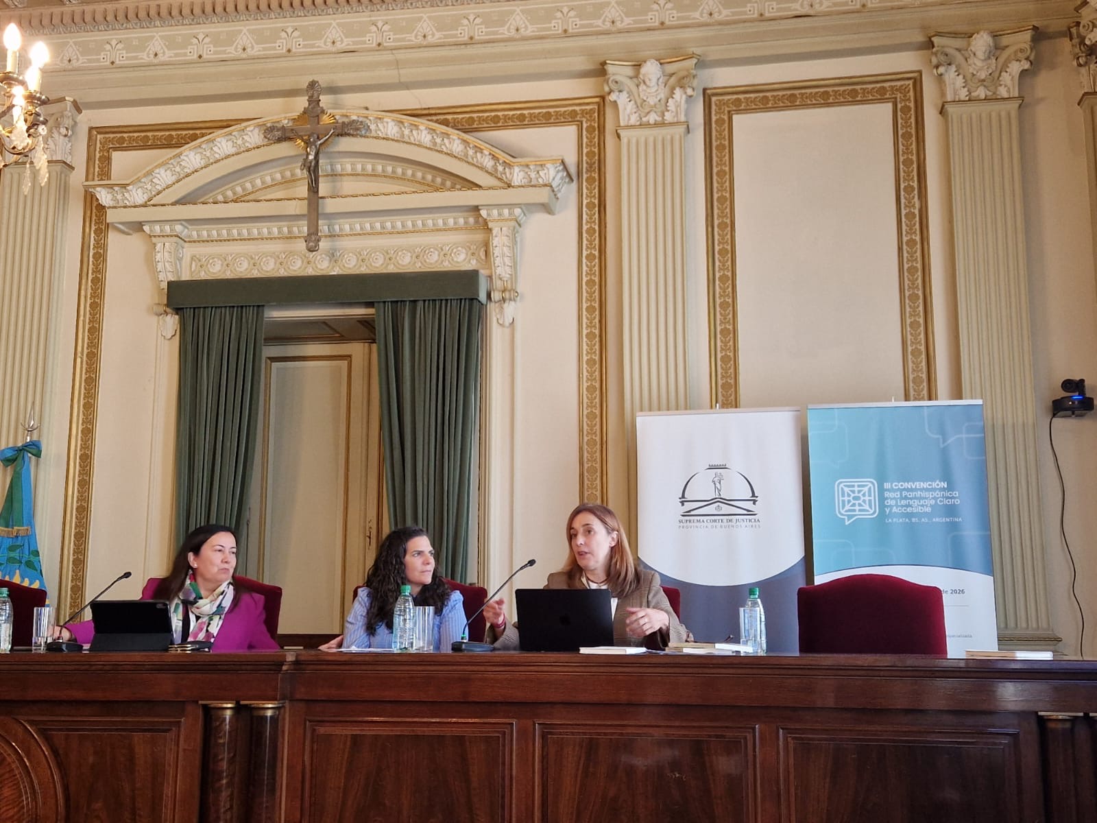 Tres ponentes en la mesa de una sala de paredes ornamentadas, bajo un crucifijo; una de ellas habla ante un portátil. A su lado, los carteles de la Suprema Corte de Justicia de la Provincia de Buenos Aires y de la III Convención de la Red Panhispánica de Lenguaje Claro y Accesible.